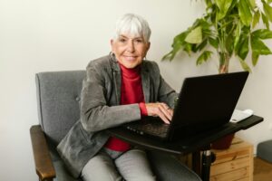 Senior adult woman in a blazer sitting at a desk with a laptop, smiling indoors.
