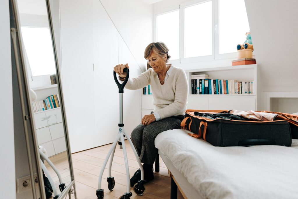 Senior woman sitting on bed with walker, preparing suitcase in bright bedroom.