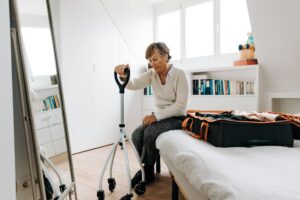Senior woman sitting on bed with walker, preparing suitcase in bright bedroom.
