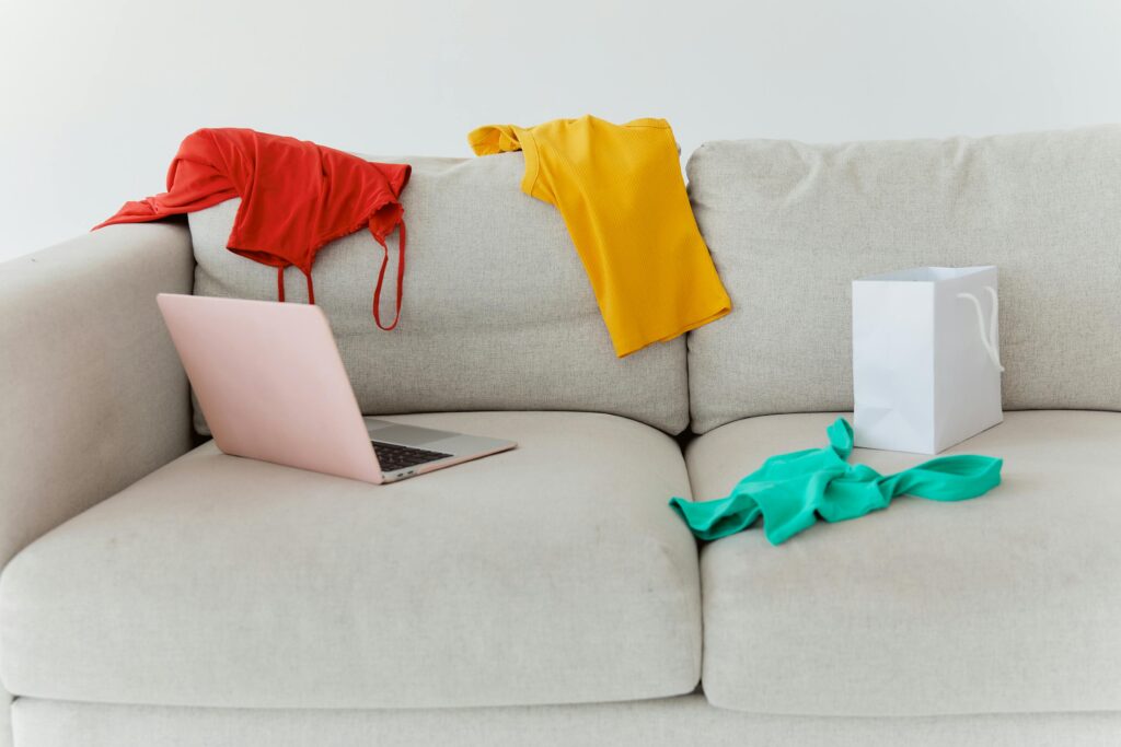 Vibrant clothes scattered on a sofa with a laptop and paper bag, showcasing a casual setting.