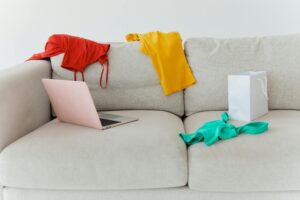 Vibrant clothes scattered on a sofa with a laptop and paper bag, showcasing a casual setting.