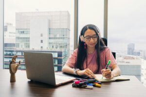 Young woman wearing headphones, writing in notebook at office desk with city view.