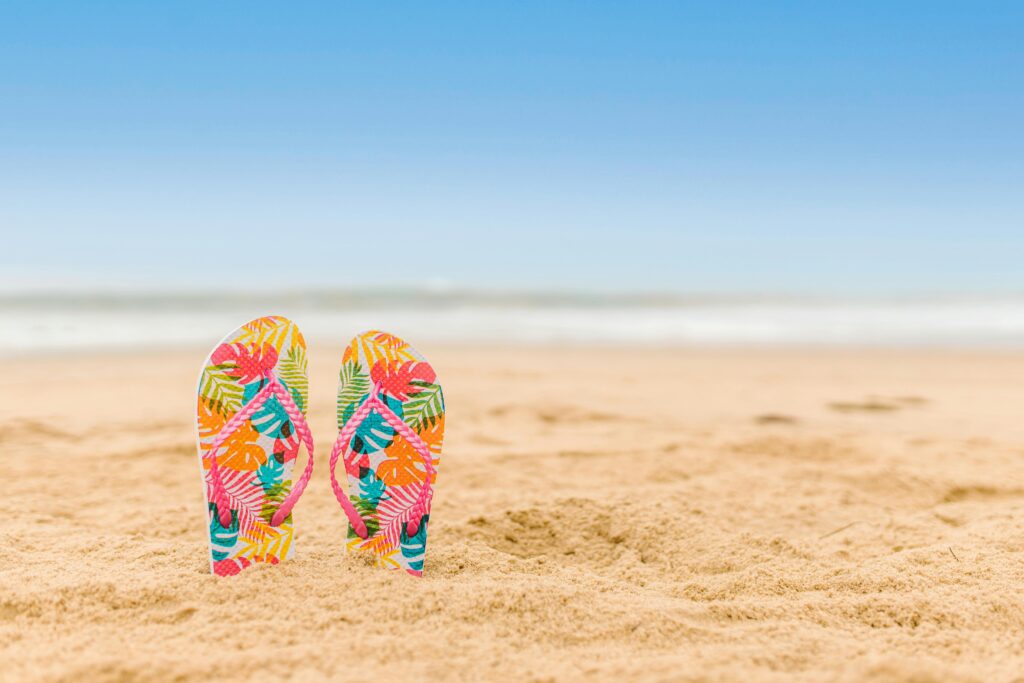 Vibrant patterned flip flops standing on a sandy beach with a clear blue sky.
