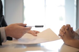 Side view of faceless formal man giving pen and paper to focused female with clenched hands at table on meeting